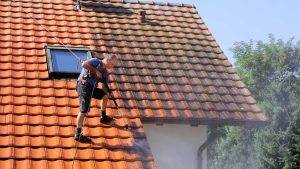 Un couvreur nettoie un toit en tuiles avec un nettoyeur haute pression, attaché par un harnais de sécurité