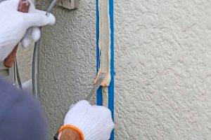 Mains gantées lissant un joint d’étanchéité le long d’une ouverture verticale, délimitée par du ruban de masquage bleu